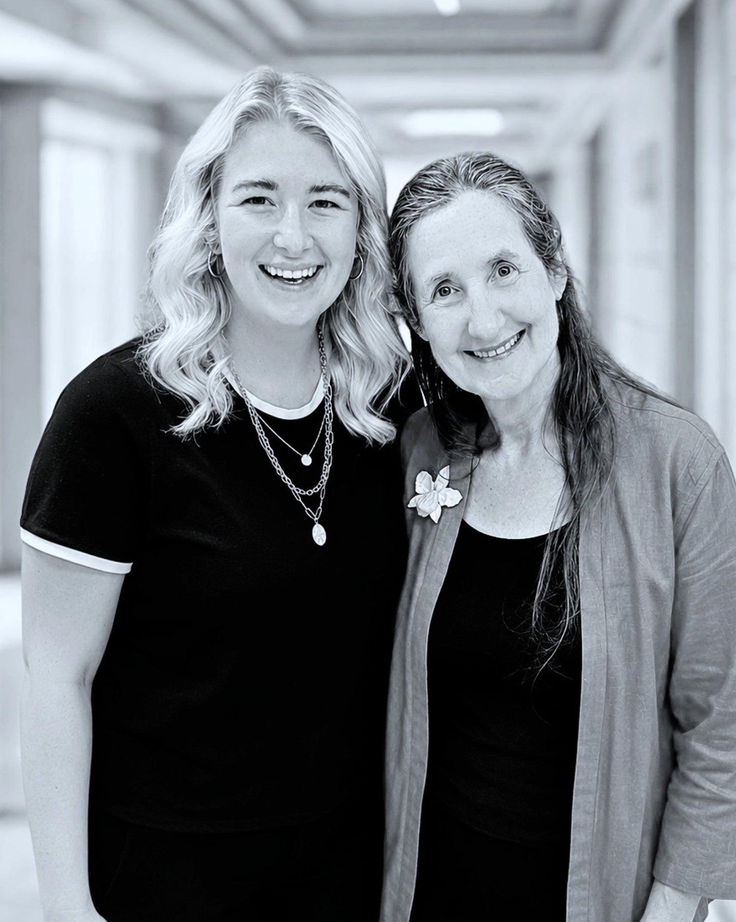 Two women standing side by side in a hallway, smiling.